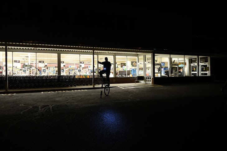 a lone mutant bicyclist rides in an empty parking lot of a closed supermarket, late at night