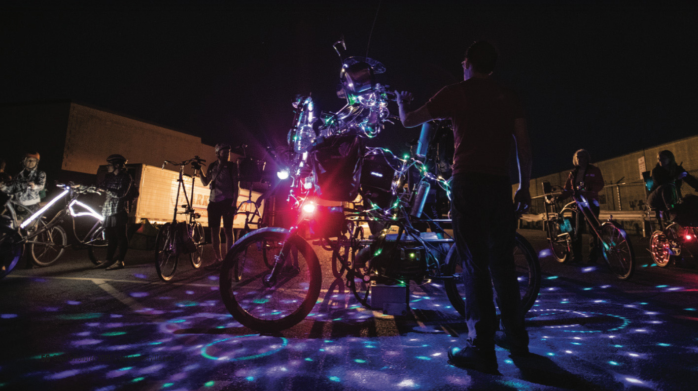 A tallbike at night with lots of lights projected on the ground, with many modified bicycles in the background, preparing for a ride.