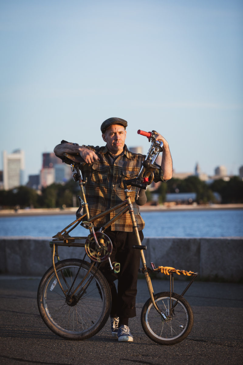 A dapper gentleman with a wool cap and plaid wool shirt poses with a tallbike with a short wheelbase, with the Boston Skyline in the background..