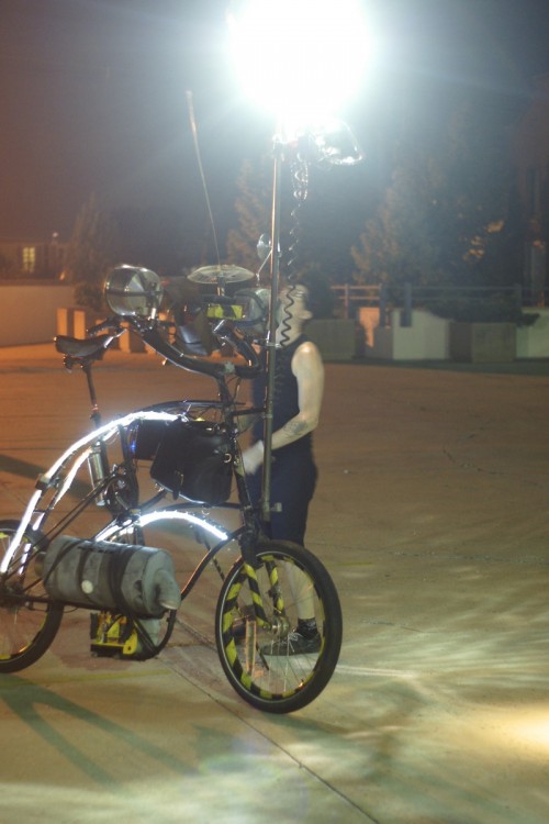 a cyclist adjusts something on his tallbike that has a brightly lit mirror ball mounted to a mast in the front.