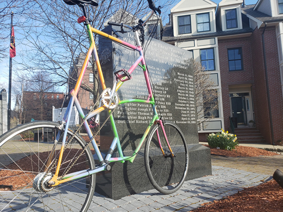 a colorful tallbike leans against a memorial to firefighters