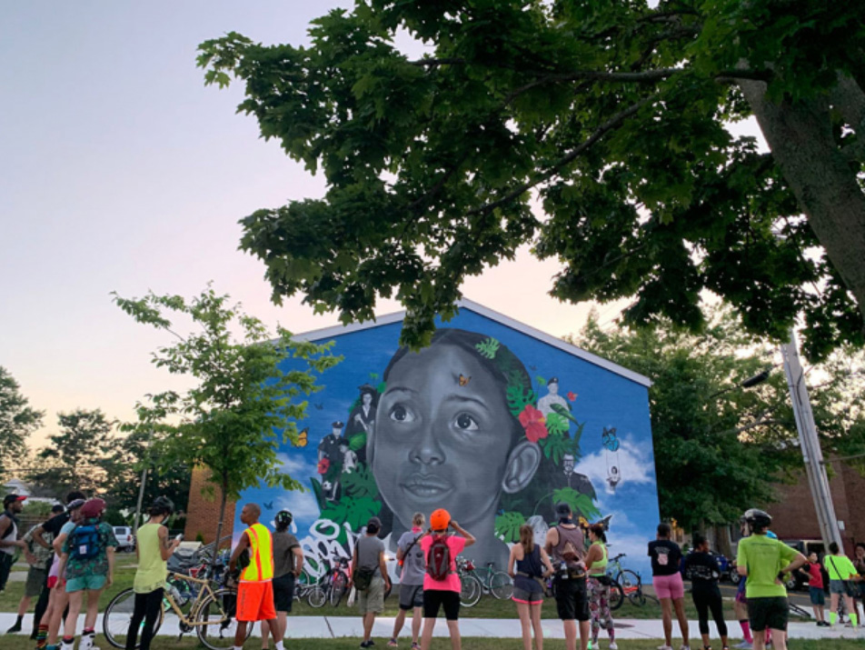 cyclists gather in front of a huge mural of a hopeful looking young girl of color